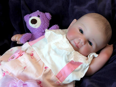 A baby doll lies on a purple blanket, accompanied by a teddy bear, evoking a sense of comfort and childhood innocence.